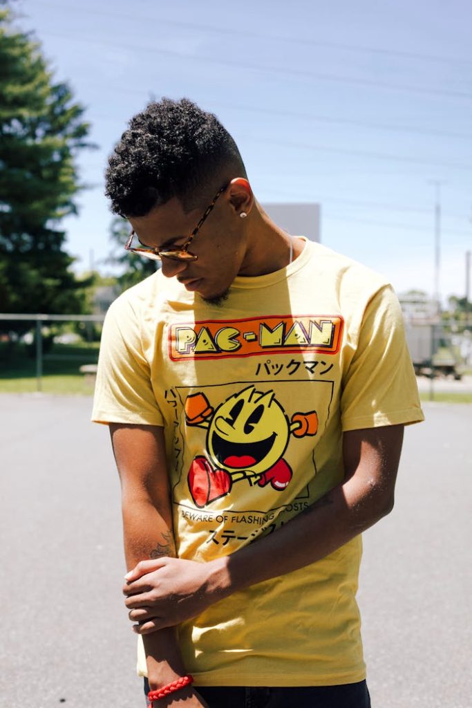 Casually dressed young man wearing a Pac-Man t-shirt, looking down outdoors under bright sunlight.